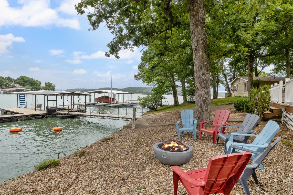 A wide view of the dock flanked by trees, Adirondack chairs by a fire pit, and sparkling water. The dock extends invitingly into the lake, showcasing both the relaxation zone (chairs/fire pit) and direct lake access.