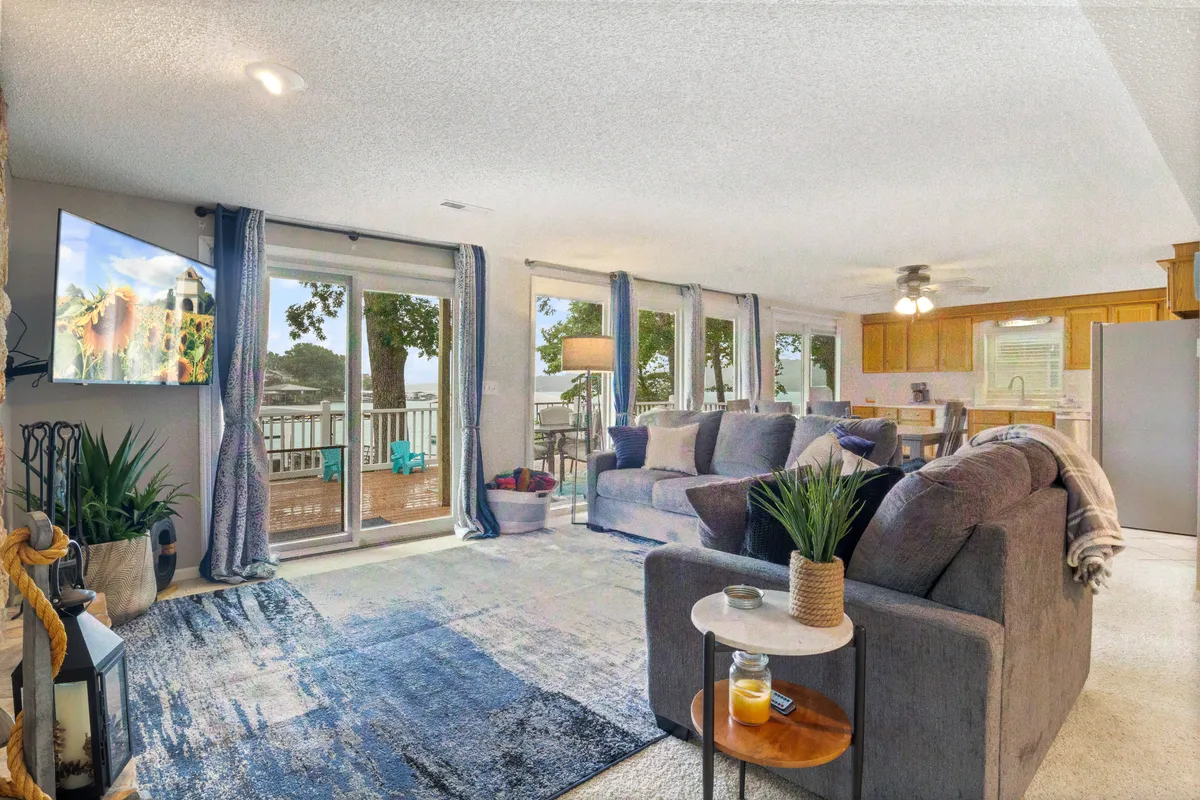 A spacious open-plan living area with a large sectional sofa facing a TV that displays a scenic landscape. Sliding glass doors lead to an outdoor space, and the kitchen area is visible in the background, creating a sense of flow between living, dining, and kitchen spaces.