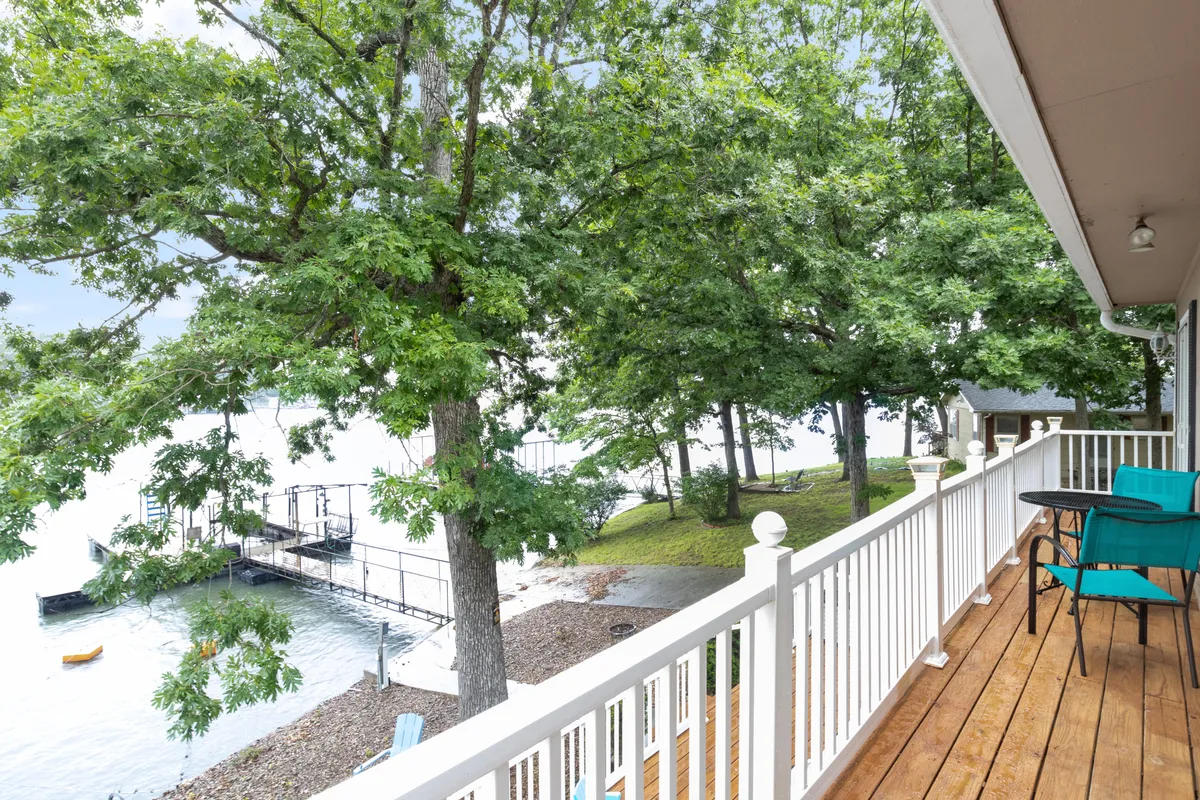 Blue chairs rest on a porch railing overlooking the lake, with green trees framing the peaceful water view. The space invites relaxation, outdoor dining, and family gatherings after a day on the lake.
