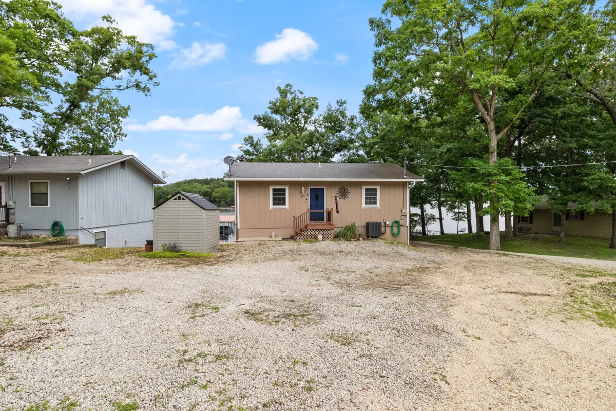 A welcoming exterior view of the main entrance featuring a distinctive blue door, surrounded by green trees and adjacent buildings. The gravel driveway and porch area suggest a relaxed, inviting environment ideal for seasonal celebrations and outdoor gatherings.