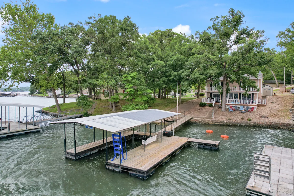 The hero image showcases the home's private dock extending over tranquil lake waters, surrounded by lush trees and neighboring houses, under a soft, colorful sunset sky. The dock is perfect for boating, fishing, or simply relaxing with friends and family, while the gentle lake ripples set a peaceful mood.