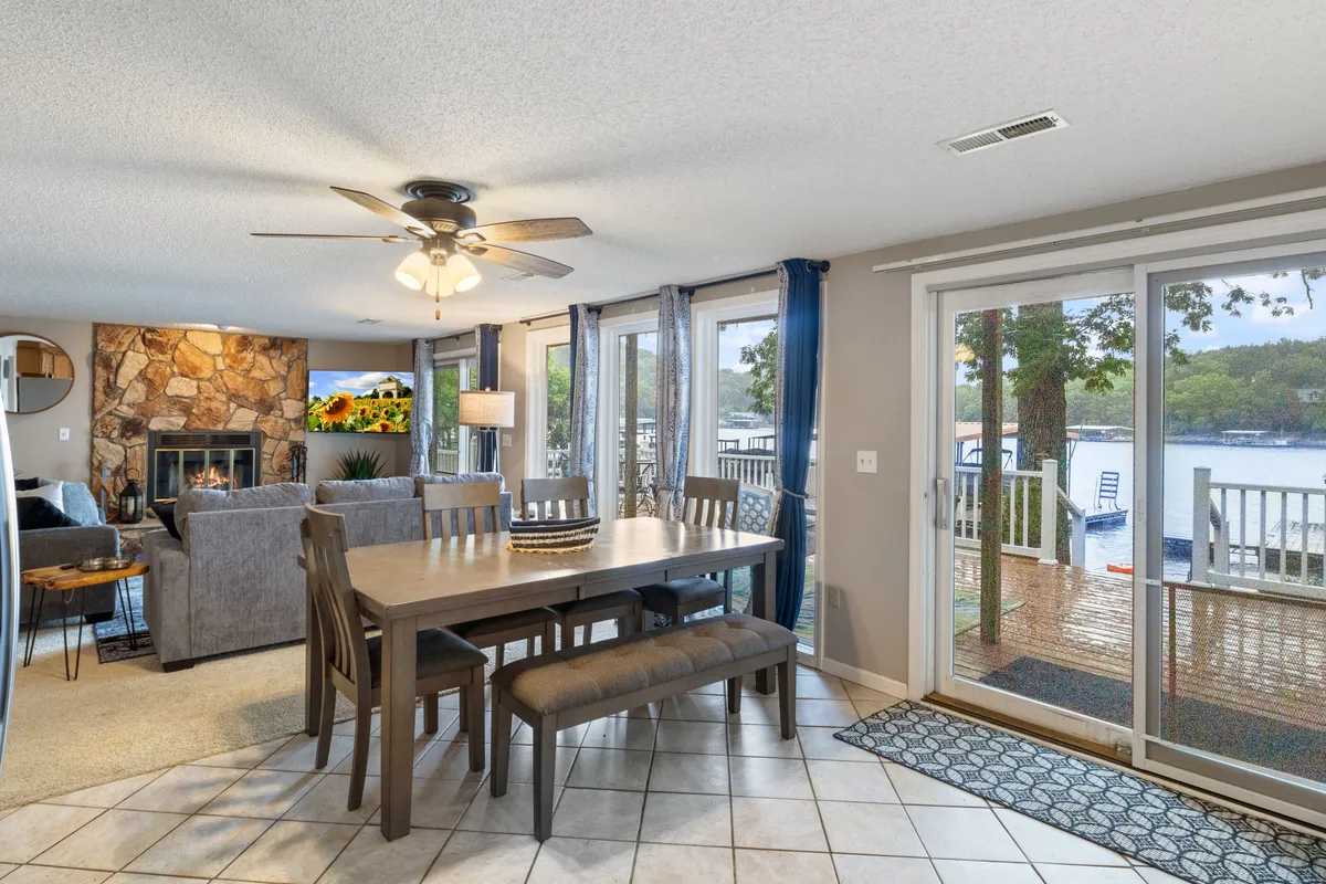 A dining table set near a cozy fireplace with large windows offering a view of the water. The arrangement invites gatherings for meals or games, with abundant natural light and a scenic backdrop.