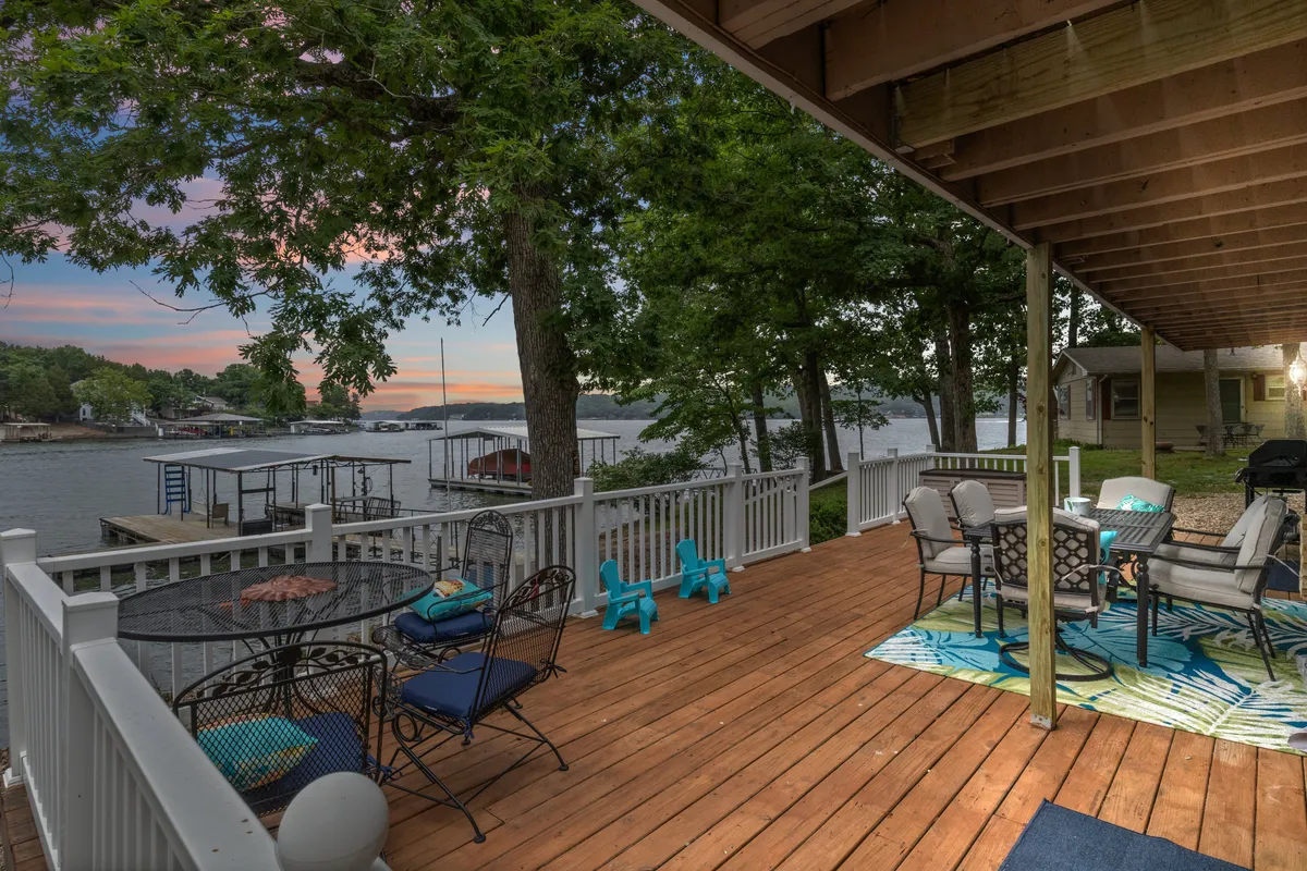 A wooden deck overlooks the shimmering water, featuring an inviting outdoor seating area with a colorful rug and a dining table set for gatherings. The deck is surrounded by lush greenery, offering tranquil lake views and a peaceful atmosphere.