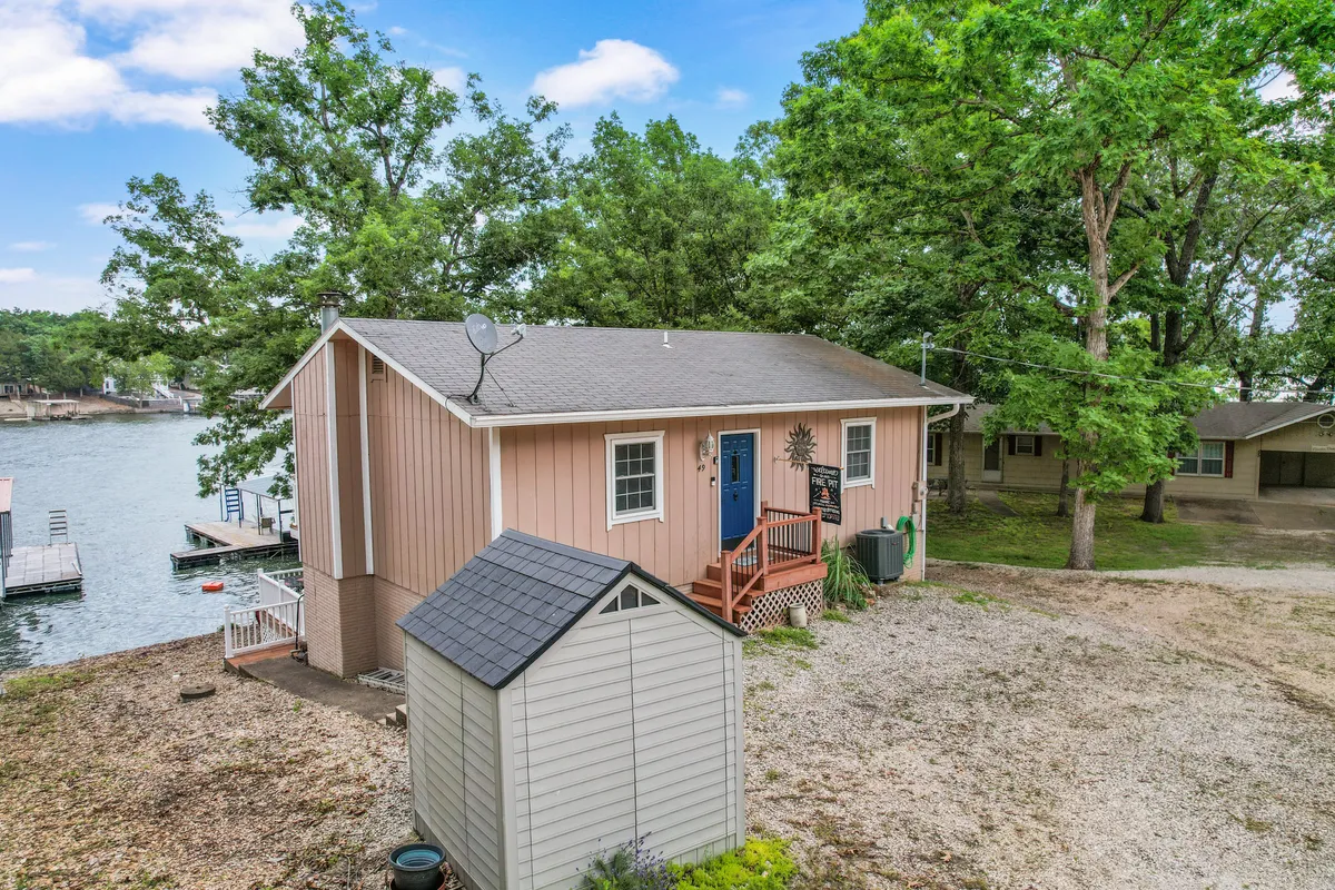 This image features the house with a blue door, a comfortable patio area, nearby water, and lush surrounding trees. The water is visible just beyond the patio, hinting at easy lake access. The warm lighting and seating invite guests to relax outdoors.