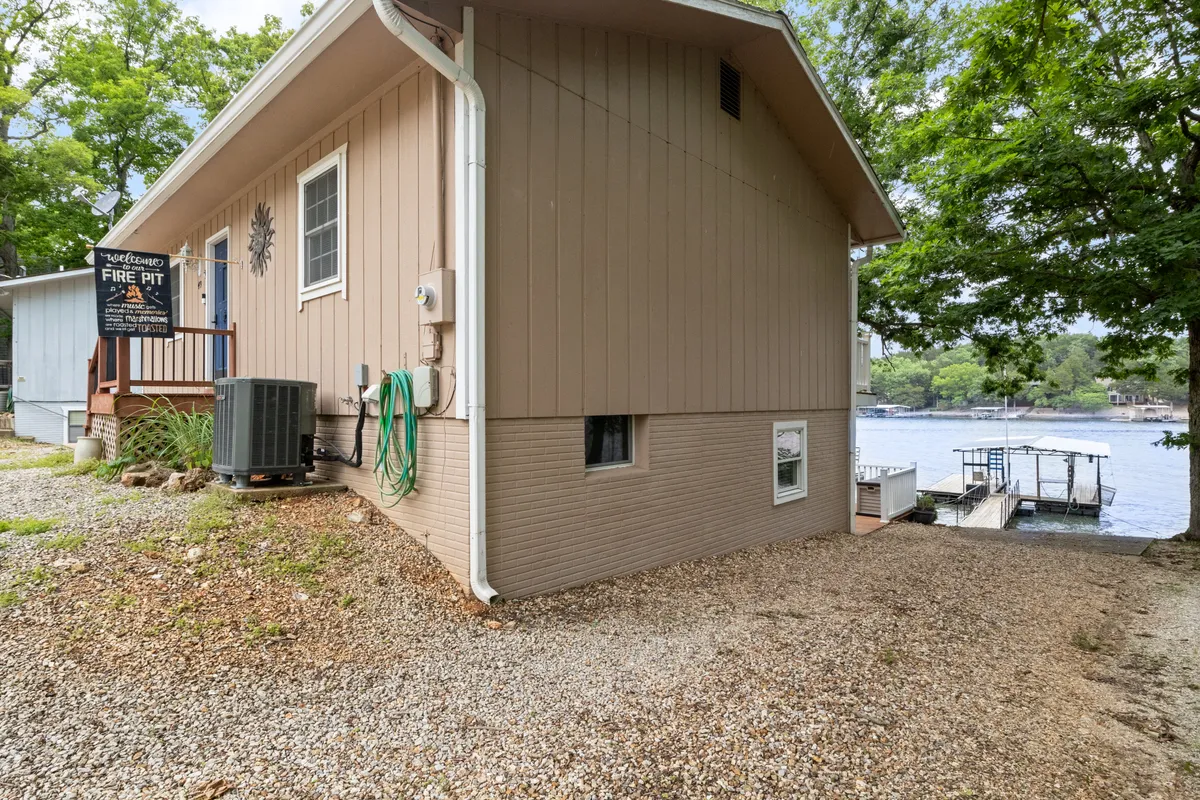 This exterior image captures a lakeside setting with a gravel pathway, lush surrounding trees, a fire pit sign, and a clear view of the lake. The composition suggests opportunities for outdoor gatherings, relaxing by the water, fishing, barbecues, and stargazing.
