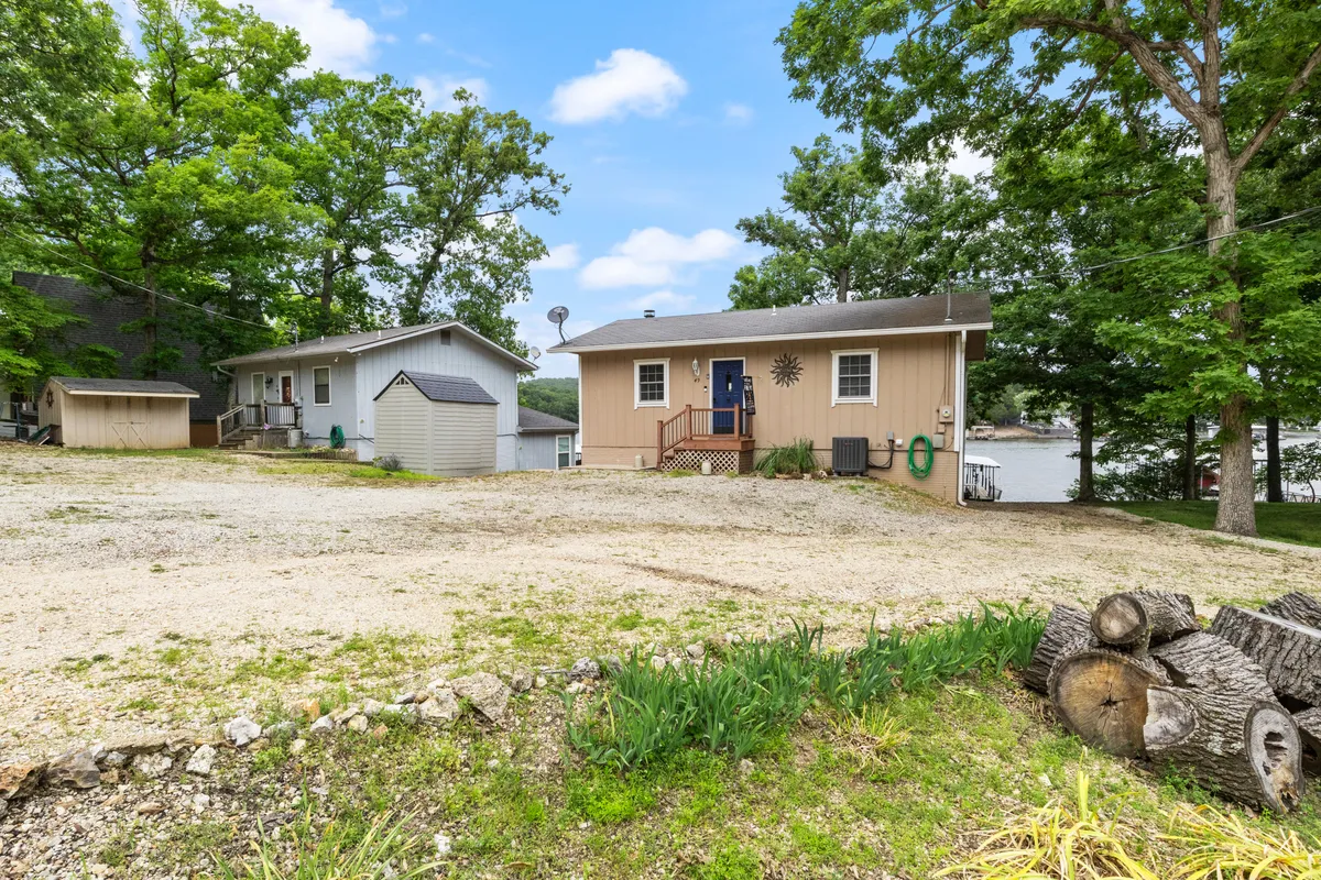 A wide exterior perspective featuring the main house, guest house, a tree-lined backdrop, and a sweeping view of the lake. The atmosphere is tranquil, evoking family gatherings, nature walks, outdoor games, and the beauty of lake sunsets and stargazing nights.