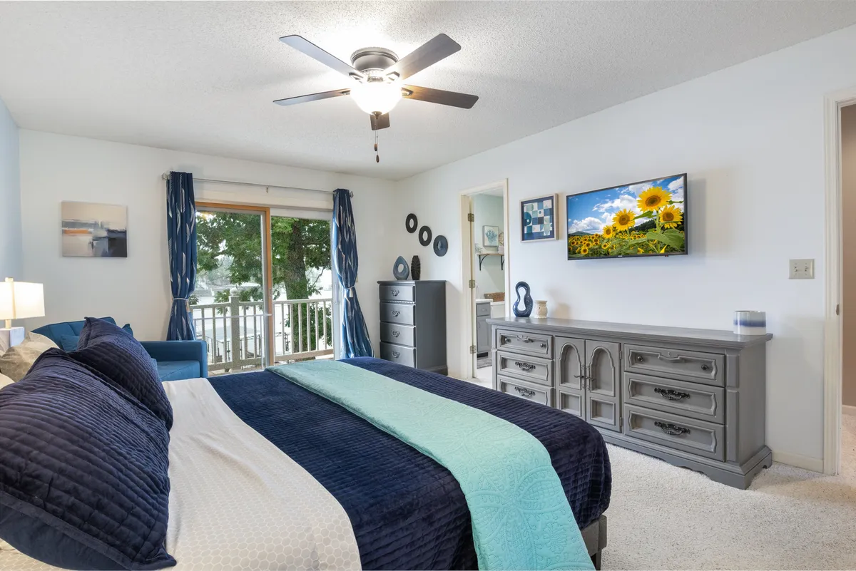A serene bedroom with navy and teal bedding, a large window leading to a balcony, wall-mounted TV, and decorative dresser. The room is filled with natural light, emphasizing the blend of comfort and access to the outdoors.