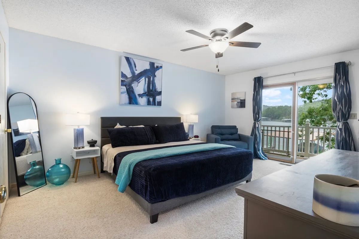 A tranquil bedroom featuring a bed with blue and black bedding, a large window offering a serene view, abstract wall art, a stylish mirror, and a blue accent table. Natural light fills the space, creating a peaceful atmosphere ideal for rest.